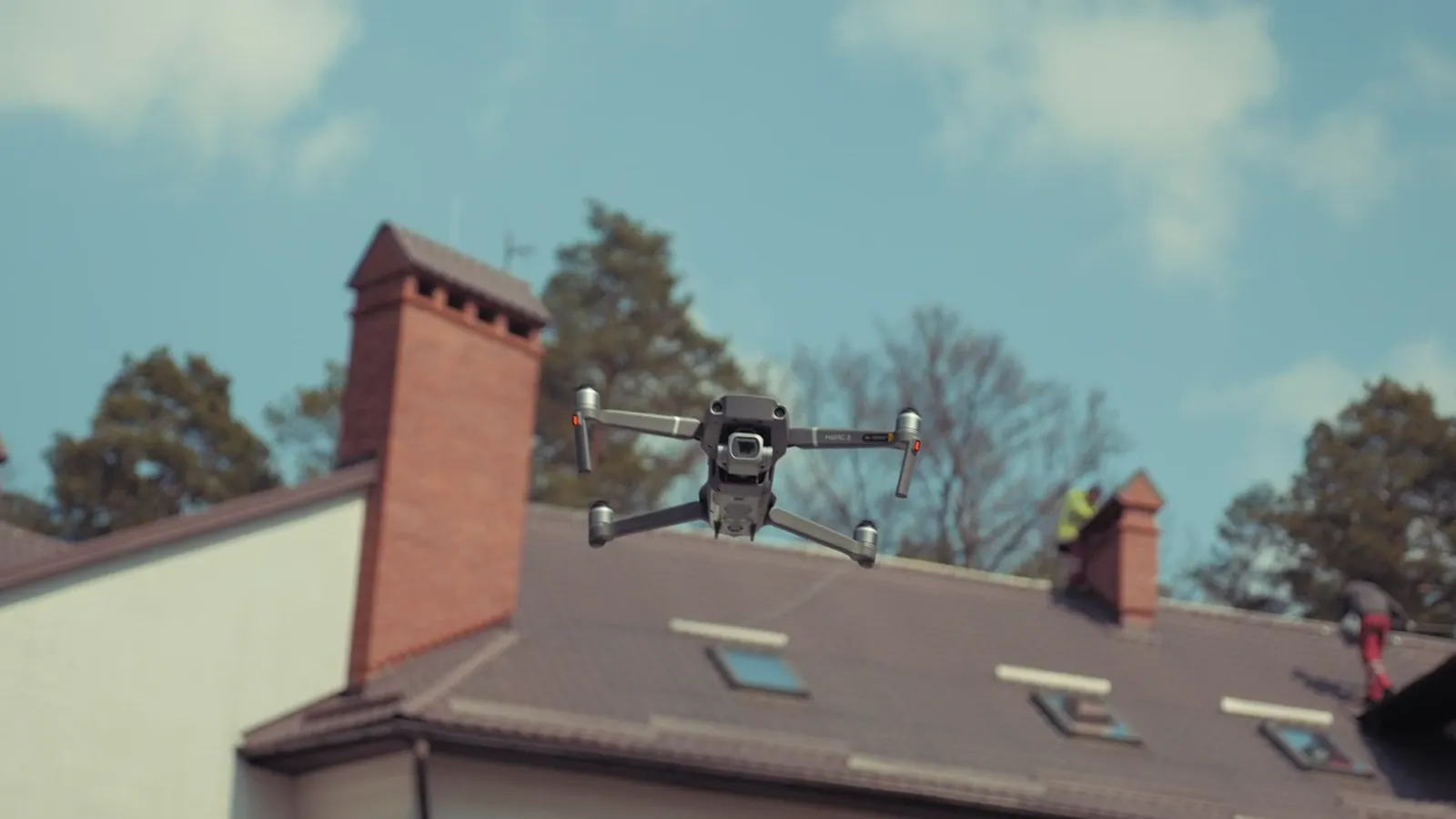 A drone inspecting a pitched shingle roof with a brick chimney and skylights